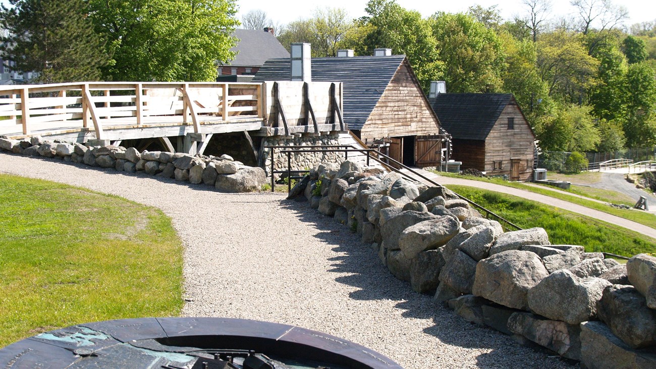 a paved path bordered by grass and stone leads to a wood bridge and two wood-sided buildings