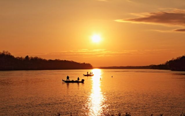 Sun setting on lake with two silhouetted boats and silhouetted trees.