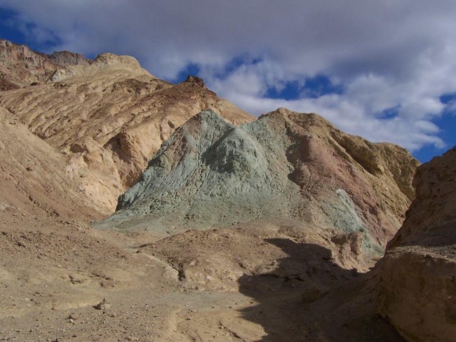 A colorful canyon with greens, blues, and yellows beneath a cloudy blue sky.