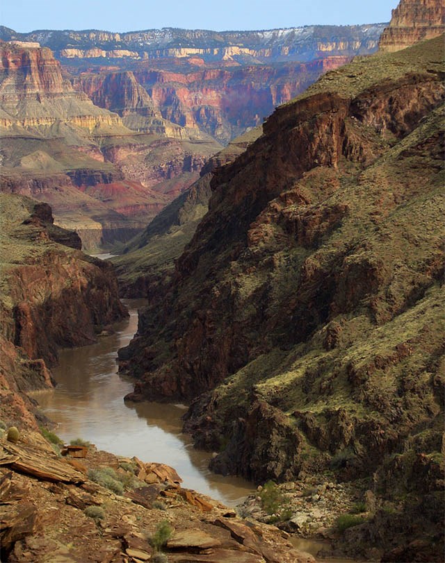 A muddy river cutting through foreground rock with a wall of stratified rock layers in the distance.