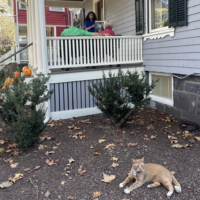 An orange cat is in the foreground with an SCA intern giving a tour on the front porch in the background.