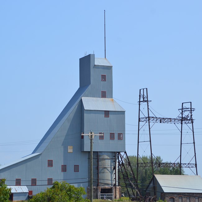 A tall metal mining structure surrounded by green foliage and a light blue sky.