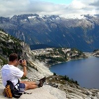 A hiker sits and photographs a mountain lake.