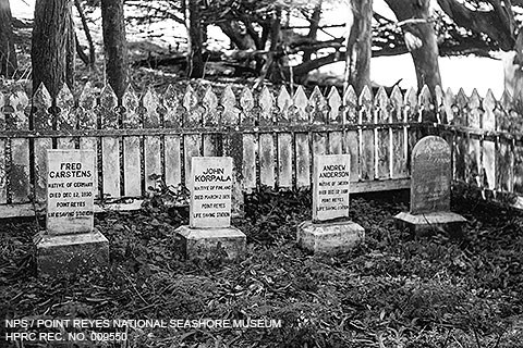 A black and white photo of four grave markers surrounded by a picket fence and trees.