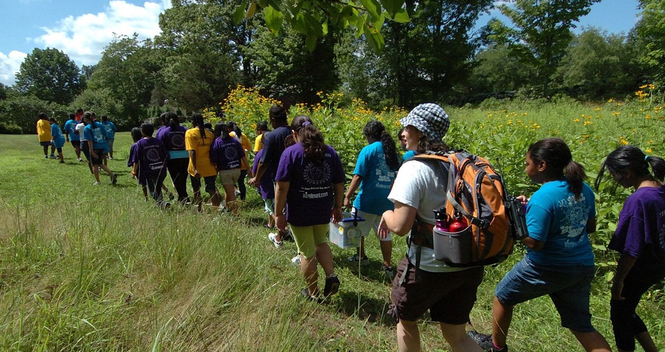 A group of school children walking in a single file line in a grassy meadow.