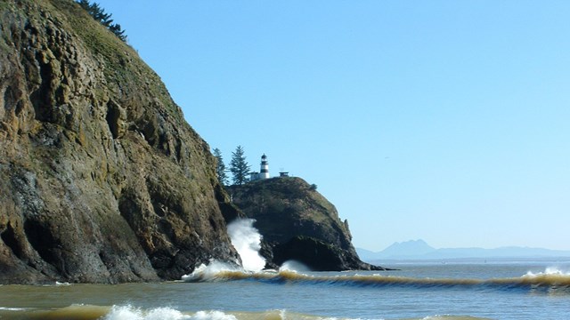 Foreground ocean waves rolling into a cove. Background high rocky bluff with lighthouse. 