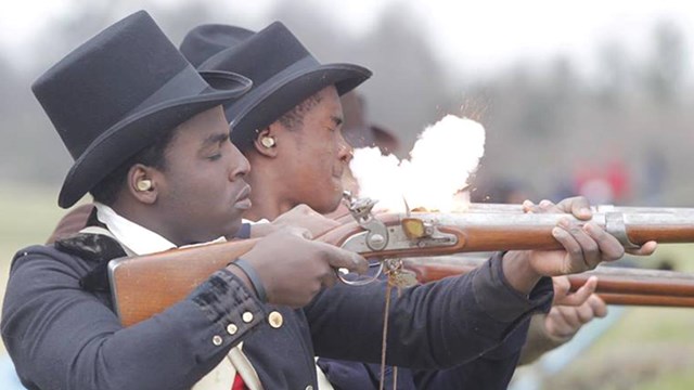 A photograph of a reinactor firing a musket, with a flash of powder