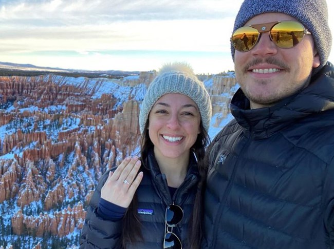 A woman holds up her hand to show a ring as she stands beside a man along a snowy cliff edge.