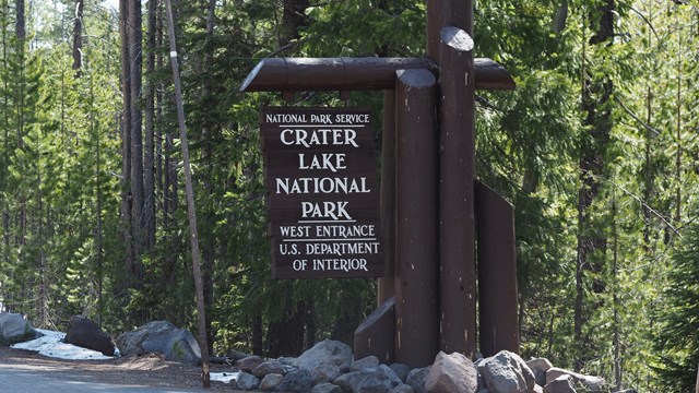 Round wood logs painted dark brown support a brown wood sign stating the west entrance to the park