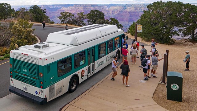 a green and white bus with passengers at a bus stop with a vast canyon landscape in the distance.