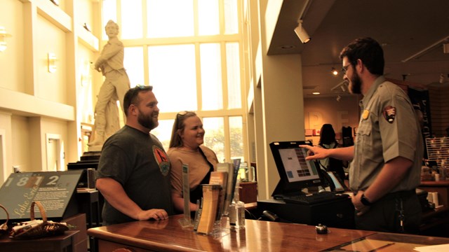 Two people stand behind a wood counter, a ranger is on the opposite side in front of a fee register