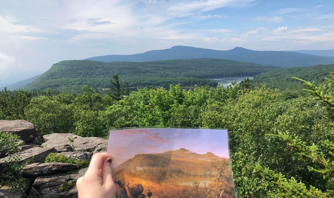 A view of a distant mountain in shades of blue & green. A hand holds a portrait image of the same. 