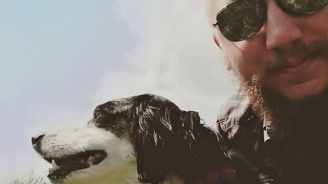 A dog wearing a harness and leash and his human wearing sunglasses take a selfie together.