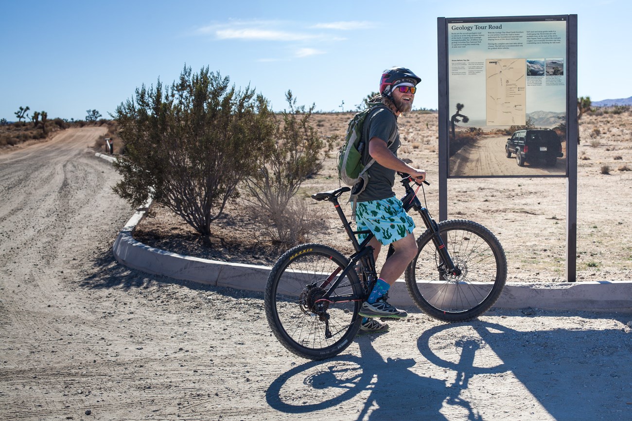 A man stands over a mountain bike at the start of a dirt road.