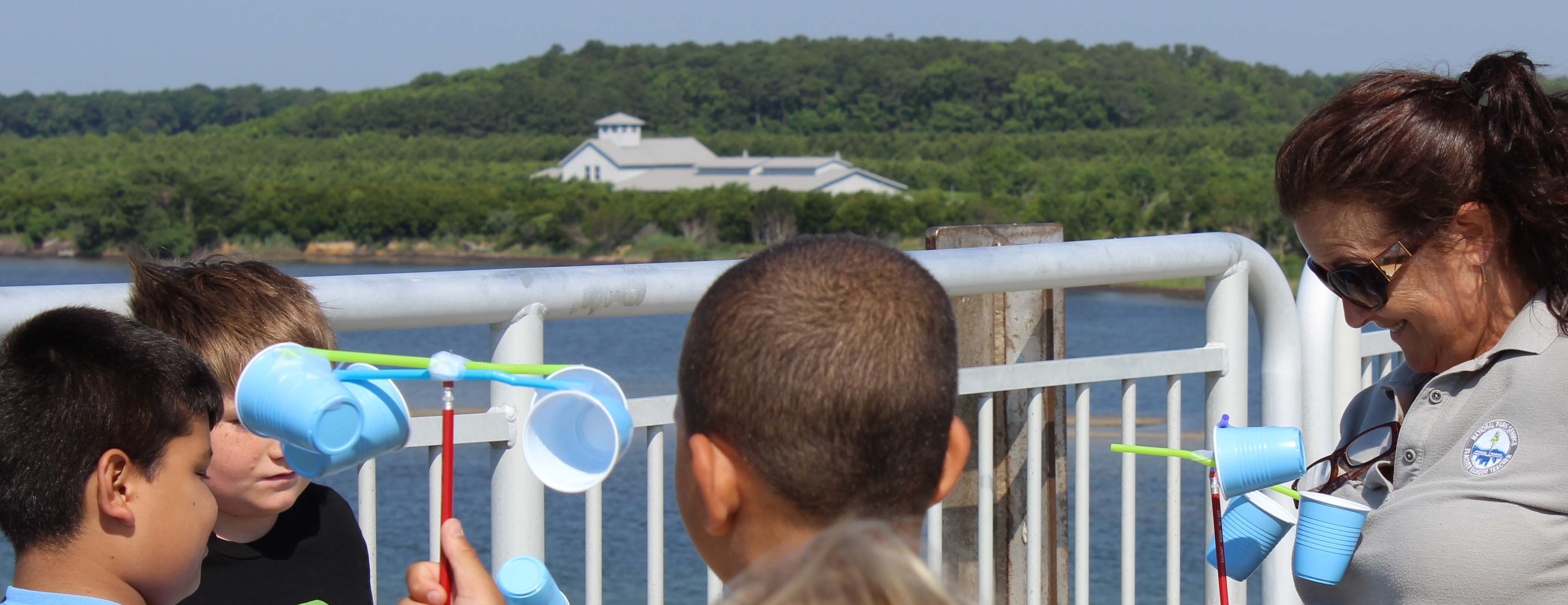 A woman and three kids hold pencils with plastic cups attached by straws next to a body of water.