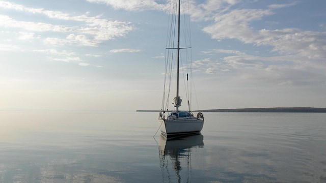 Sailboat with sails down resting on calm water with the sky and clouds reflecting off the surface. 