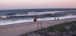 Couple strolls down an empty beach past an unstaffed lifeguard stand, drenched in a pink glow from the sunset to the west.