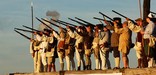 Soldiers stand on the walls of the fort at sunset, muskets leveled at an unseen target. Grey smoke lingers in clumps surrounding them.