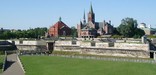 Blue skies above, a green lawn below, in between a wooden fort rises, with old churches and red brick city buildings even taller behind.