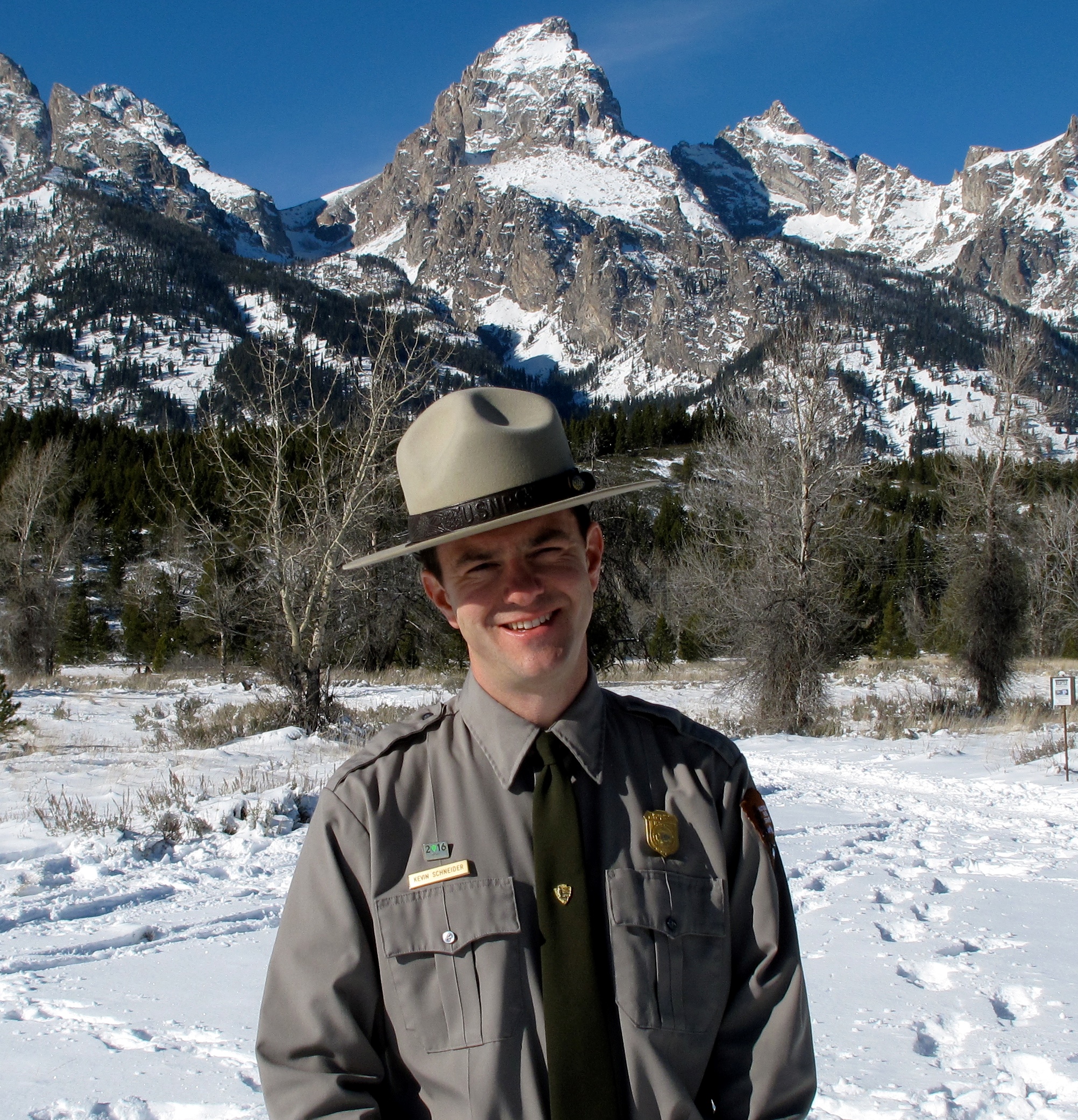 A man wearing a ranger uniform standing in front of snow covered rocky mountain peaks.