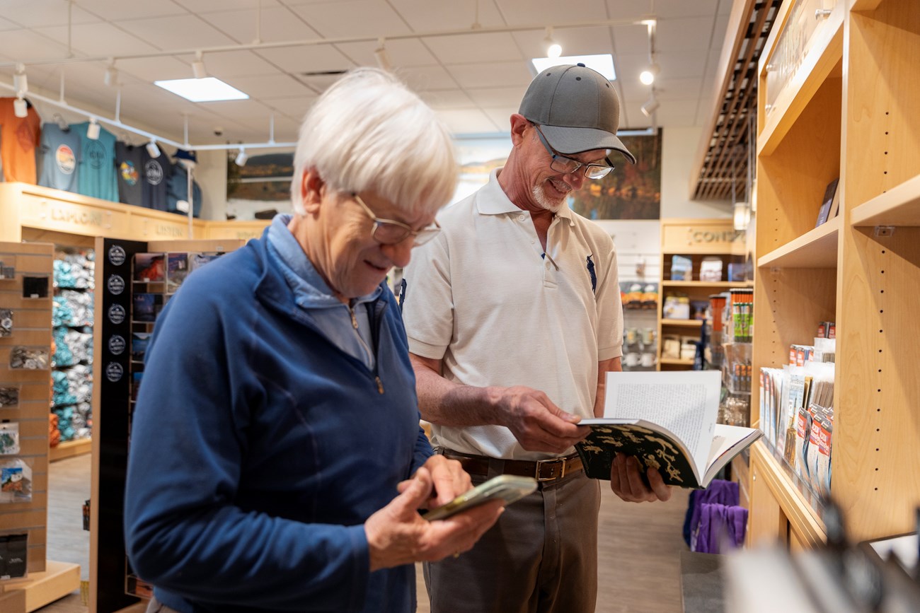 Two older adults exploring merchandise in Acadia's Park Store, with the woman examining her phone and the man reading a book, amid shelving filled with souvenirs.