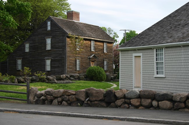 Pictured are two New England "saltbox" style homes. On the left, the home has brown wooden siding. On the right, only a portion of a home with gray-siding.