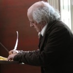 An elderly white man with graying hair wearing 1700s-style clothing writes with a quill pen at a desk