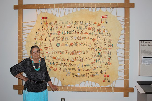 Woman stands in front of a flat, painted elk hide hung on a wooden frame & mounted to the wall. 5 spirals of fist-sized pictures appear on the hide.