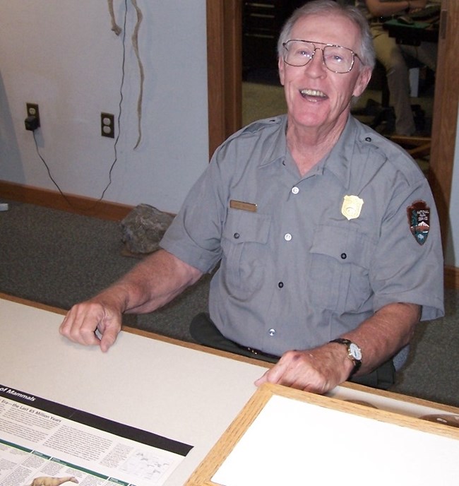 Man with short gray hair and wire-rimmed glasses sits behind a low desk and shines a big smile at us. A flyer with Agate fossils rests on the desk.