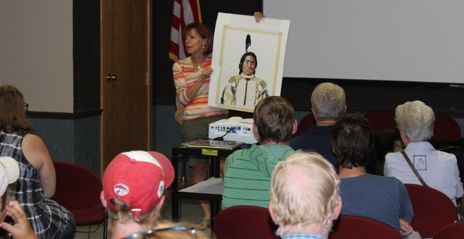 A woman holds up a painting of a native American man in front of a group of people sitting down.