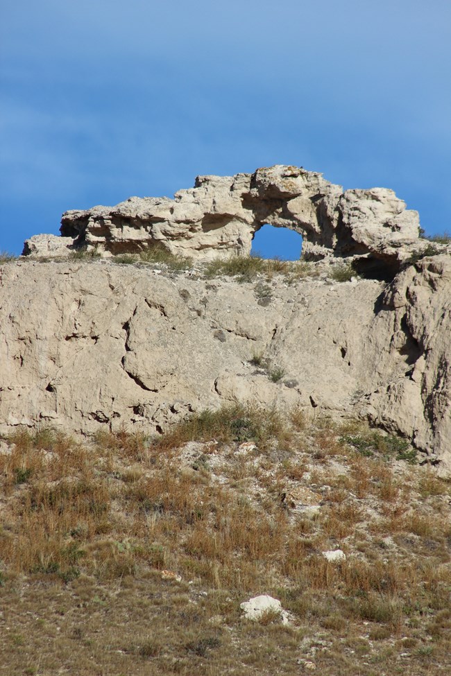 Blue sky peaks through a hole carved in a large rock that sits on top of a concave, tan boulder with prairie grass in the foreground.