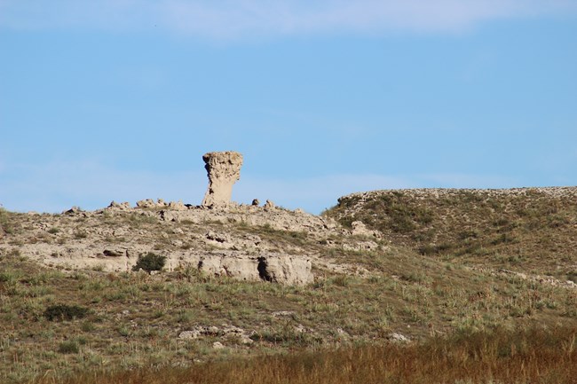 A rectangular portion of tan rock with a thicker top sticks up from a rocky hill with prairie grass in the foreground.