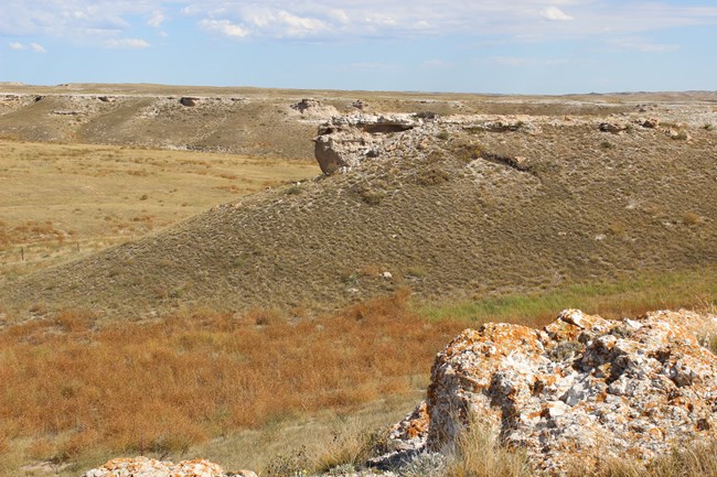 Tan colored boulders spattered with orange splotches jut out from several places on the top of a curved hill overlooking a valley of brown prairie grass.