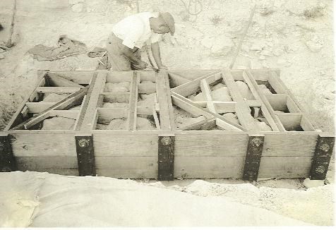 An old black and white picture of a worker with a cap building a wooden crate around a slab of rock.