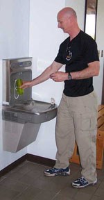 Man in black shirt and brown pants holding water bottle under a metal dispenser.