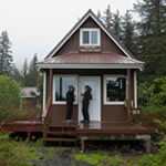 Small brown-colored cabin with porch in a wooded setting. Two people standing in front of its white door.