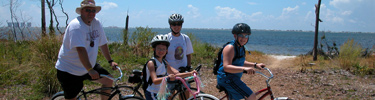 Family of four waiting to ride their bikes are in front of a trail to the blue water.