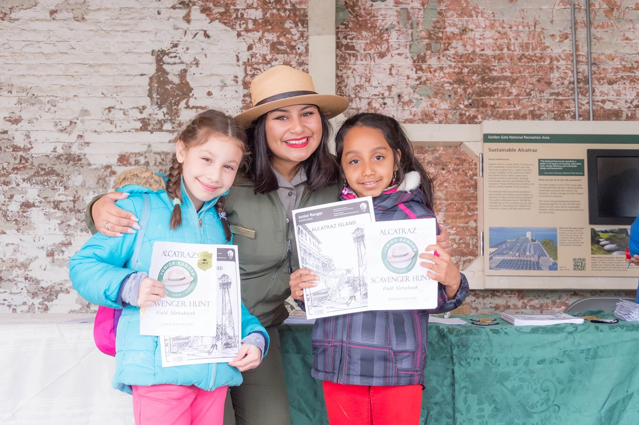 Everyone is welcome to become a Junior Ranger. Park Ranger stands between two girls holding Junior Ranger Books.