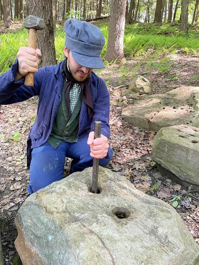 A park ranger in 19th century clothing uses tools to cut stone.