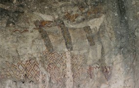 Three brown and white anthropomorphs, or humanoid images, with red hashing beneath on the wall of a cave.