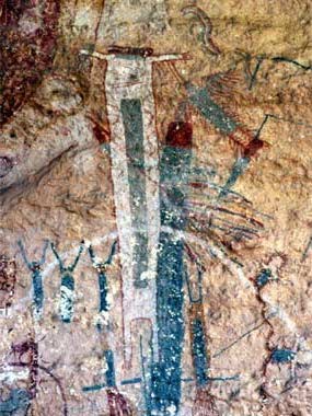 Cave painting depicting several very small, anthropomorphs with their hands over their heads to the left of a larger, white with blue rectangle in center of body, anthropomorph and a blue anthropomorph with red head and feathers.