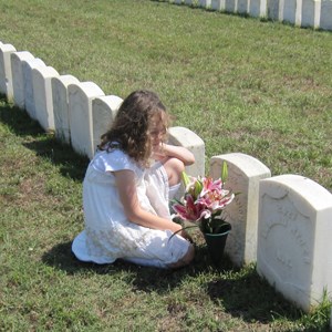 Young girl kneeling next to a grave decorated with flowers.