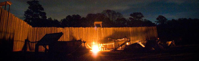 A campfire illuminates replica shelters and wooden prison walls.