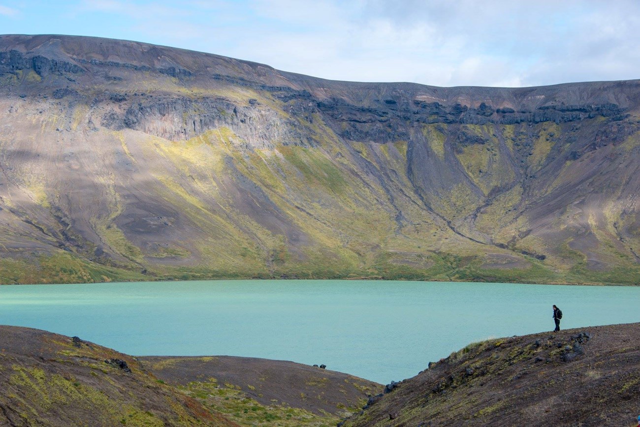 Silhouette of a hiker in the distance with a blue lake and tall caldera rim in the background