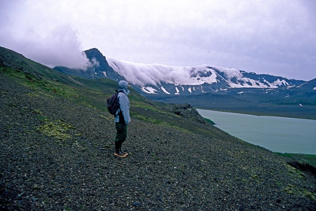 A hiker stands with rain gear while clouds roll in over caldera rim in background