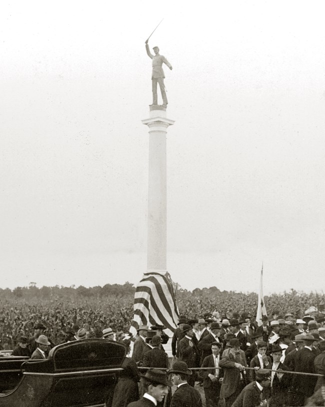 monument with soldier at the top of a column american flag draped around the base gathering of people in black and white photo