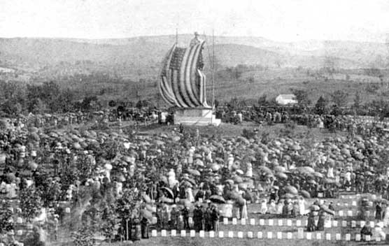 Black and white photo of a large crowd gathered in a field around the Private Soldier Statue. A huge US flag is draped over the statue and is blowing in the breeze.