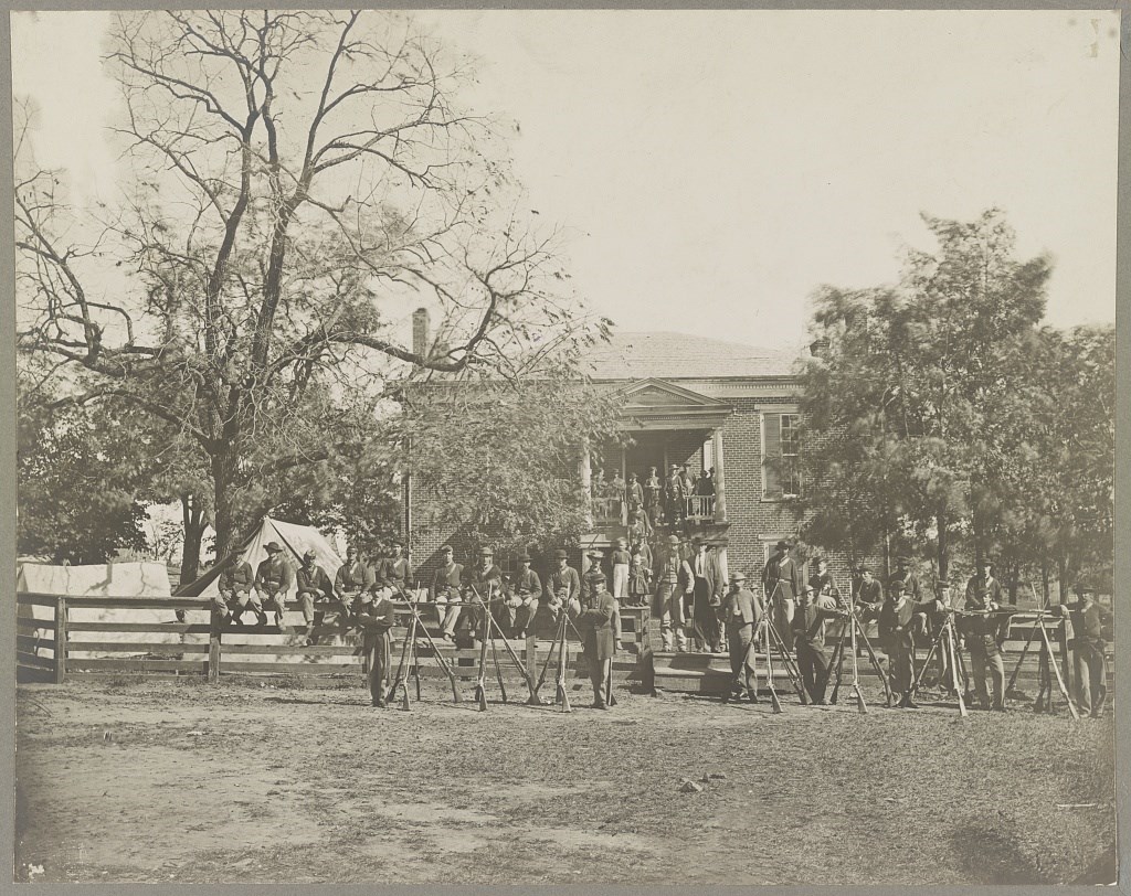 black and white photo of soldiers lined up in uniform outside a 2 story brick building, some of the men are standing, some are sitting on railings and steps