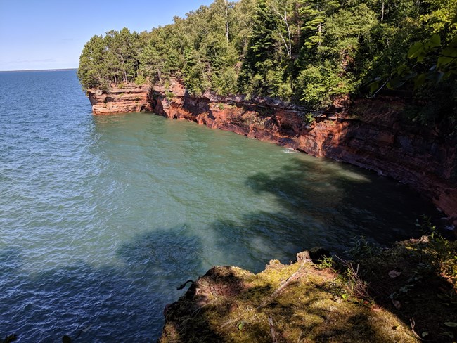Tree-covered red cliffs meet greenish blue water, in a bowl shape.
