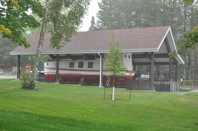 A white and red fish tug sits under a pavilion on the other side of a green grassy lawn.